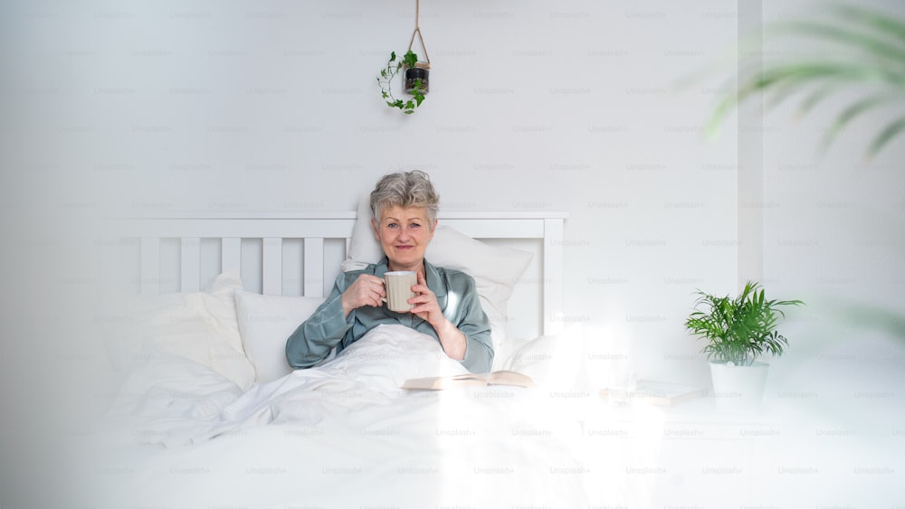 Porträt einer glücklichen älteren Frau mit Kaffeelesebuch im Bett zu Hause, Blick in die Kamera.