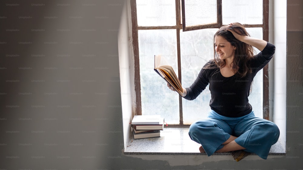 Retrato de mujer feliz sentada en el alféizar de la ventana vieja y sucia, leyendo un libro.
