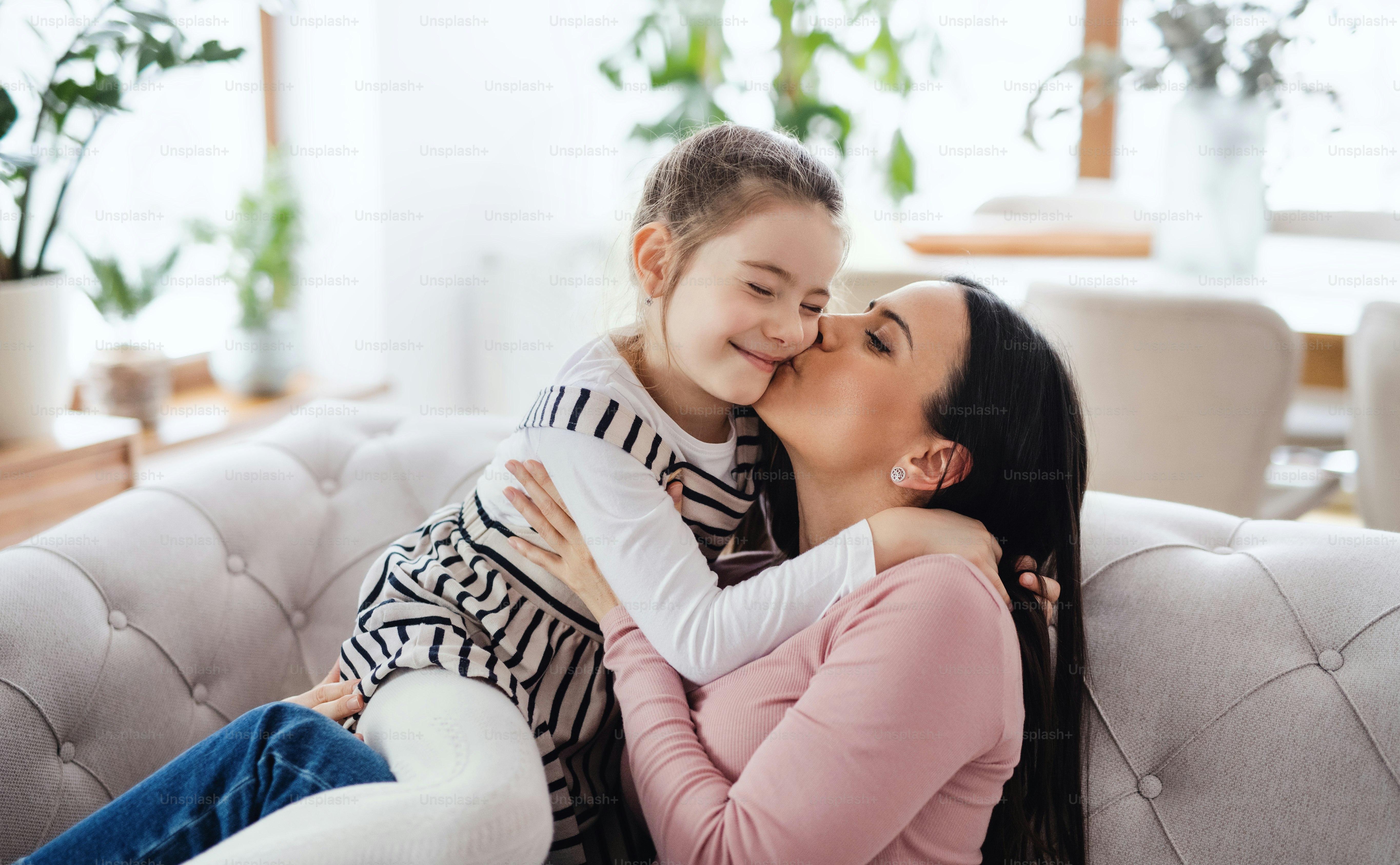 Potret ibu dengan anak perempuan kecil duduk di sofa di dalam ruangan di rumah, berciuman.