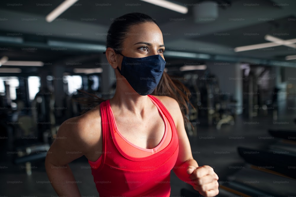 Side view of woman with face mask doing exercise on treadmill in gym, coronavirus concept.