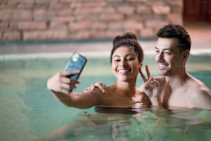 Ritratto di giovane coppia felice nella piscina termale termale coperta, scattando selfie.