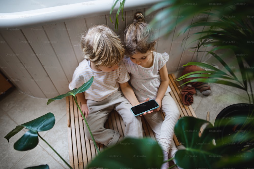 Top view of cute small children sitting on floor indoors at bathroom, using a smartphone.