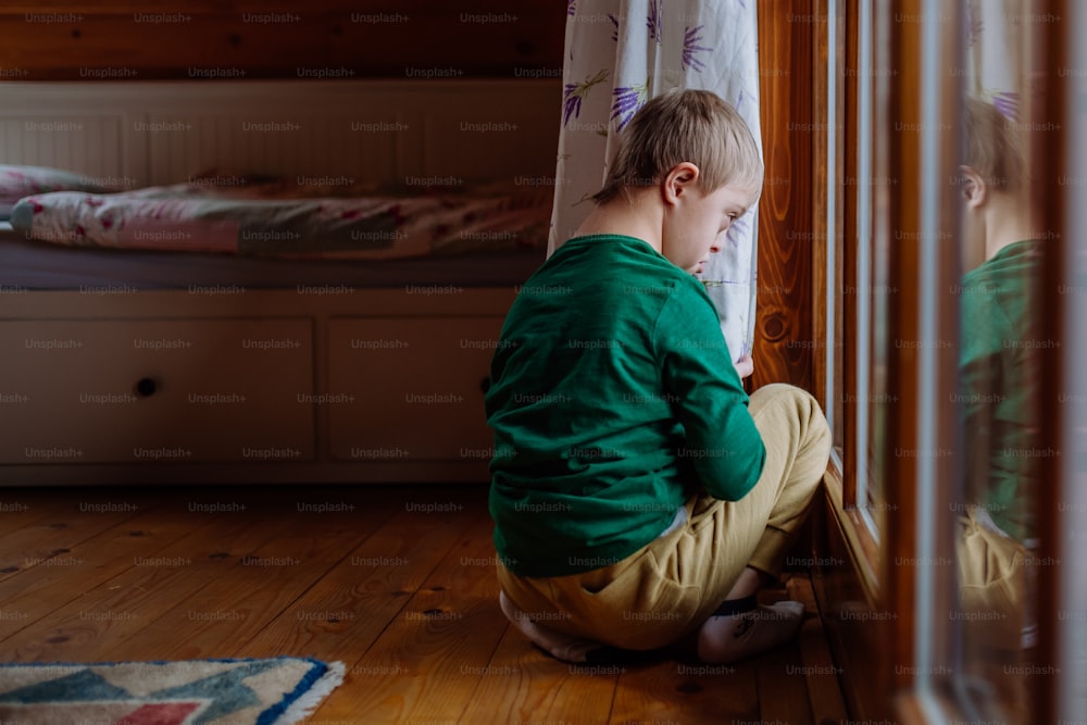 Un niño pequeño con síndrome de Down sentado en el suelo y mirando a través de la ventana de su casa.