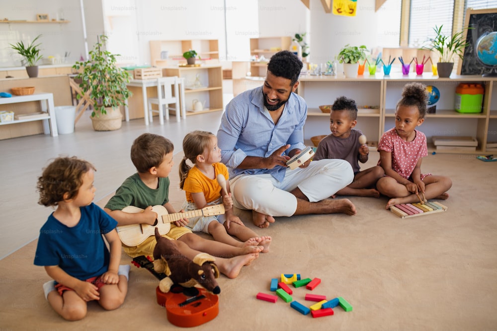 Um grupo de crianças de uma creche pequena com um professor sentado no chão dentro de casa em sala de aula, tocando instrumentos musicais.