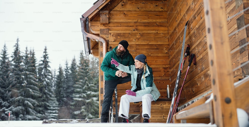Mature couple resting by a wooden hut outdoors in winter nature, cross country skiing.