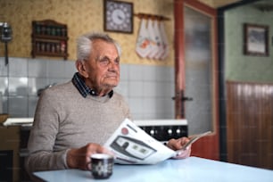 A portrait of elderly man sitting at the table indoors at home, reading newspapers.