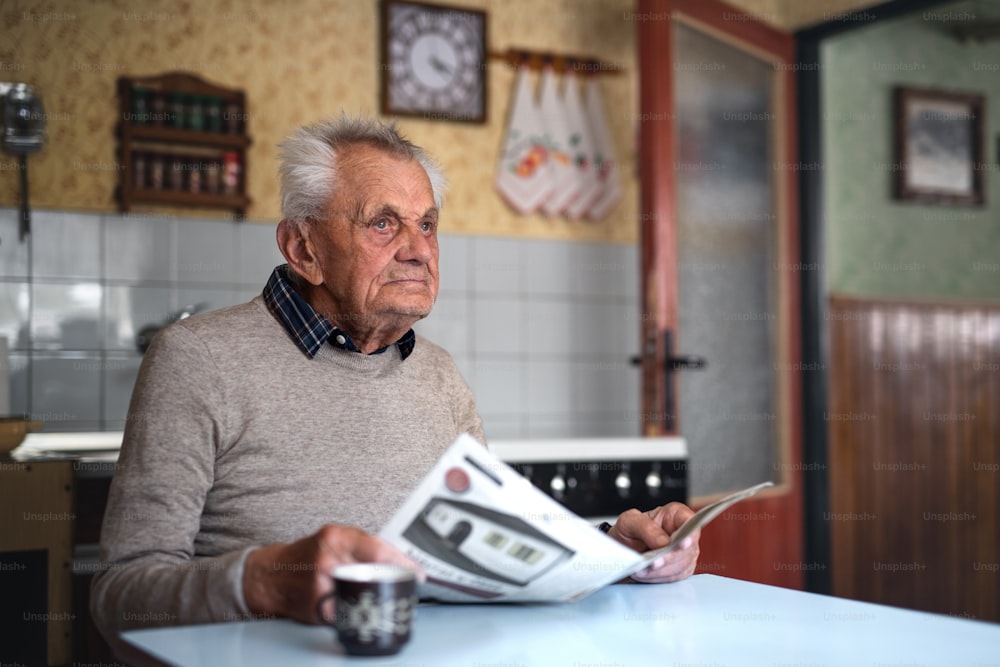A portrait of elderly man sitting at the table indoors at home, reading newspapers.