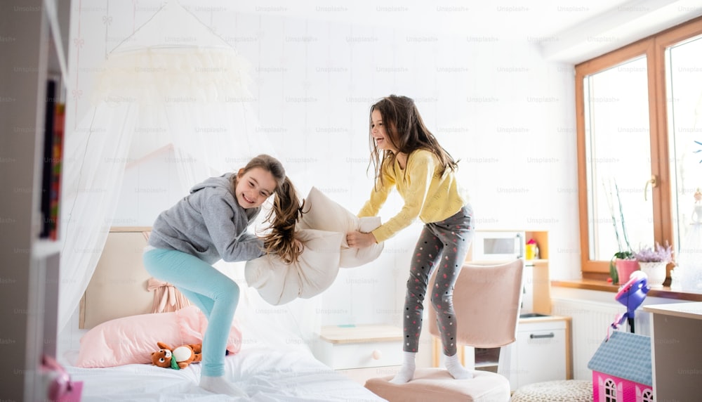 Duas alegres irmãs pequenas dentro de casa, brigam de travesseiro na cama no quarto.