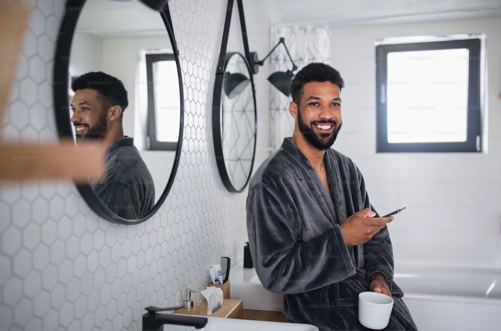 Ein junger Mann mit Kaffee und Bademantel drinnen im Badezimmer zu Hause, Morgenroutinekonzept.