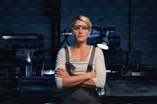 A portrait of mid adult woman standing with arms crossed indoors in metal workshop, looking at camera.