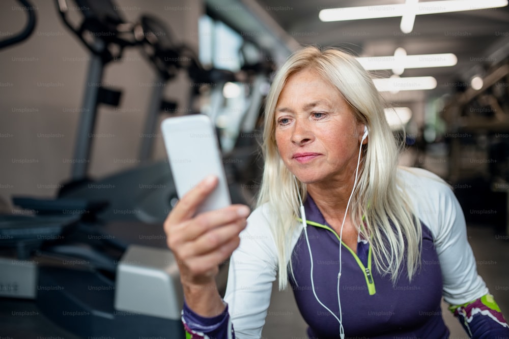 Une vue de face d’une femme âgée avec des écouteurs et un smartphone dans une salle de sport se reposant après avoir fait de l’exercice.