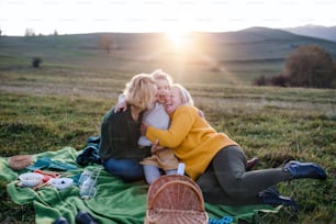 Happy small girl with mother and grandmother having picnic in nature at sunset, having fun.