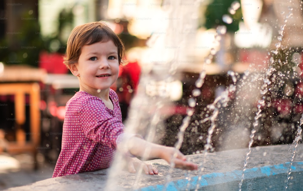 Portrait of small girl standing by water fountain outdoors in town.