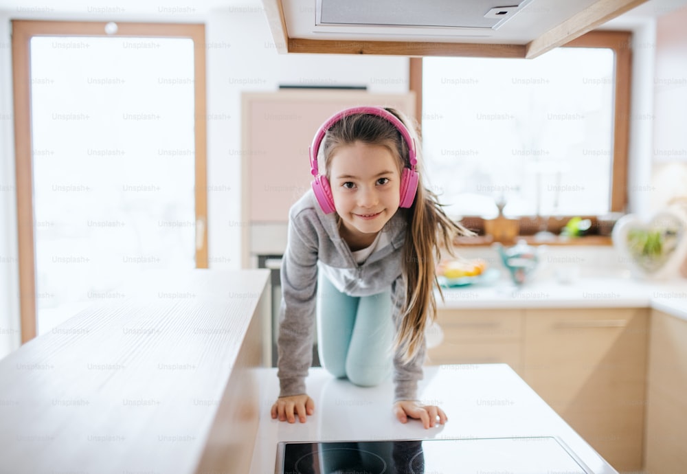 Retrato de una niña pequeña con auriculares en el interior de la encimera de la cocina en casa, mirando a la cámara.