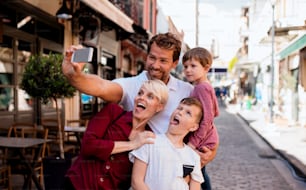 Una familia joven con dos niños pequeños parados al aire libre en la ciudad, tomándose selfies con un teléfono inteligente.