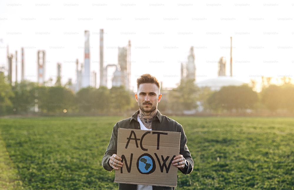 Portrait of young activist with placard standing outdoors by oil refinery, protesting.