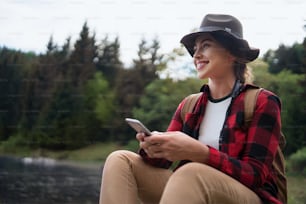 Side view of young woman sitting by lake on a walk outdoors in summer nature, using telephone.