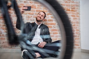 Un jeune homme d’affaires avec un vélo et un téléphone assis par terre dans un bureau, en train de prendre un selfie.