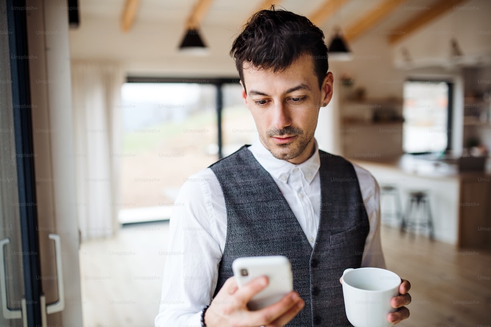 Un joven con café y teléfono inteligente parado en el interior de su casa.