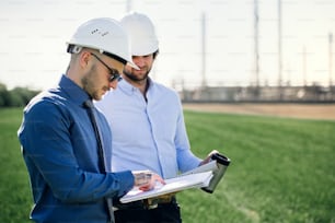 Two young engineers with hard hat standing outdoors by oil refinery, discussing issues. Copy space.