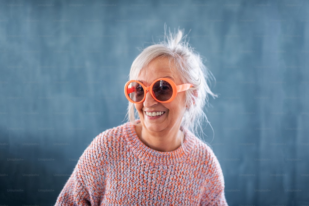 Portrait of senior woman with eccentric sunglasses standing indoors against dark background, laughing.