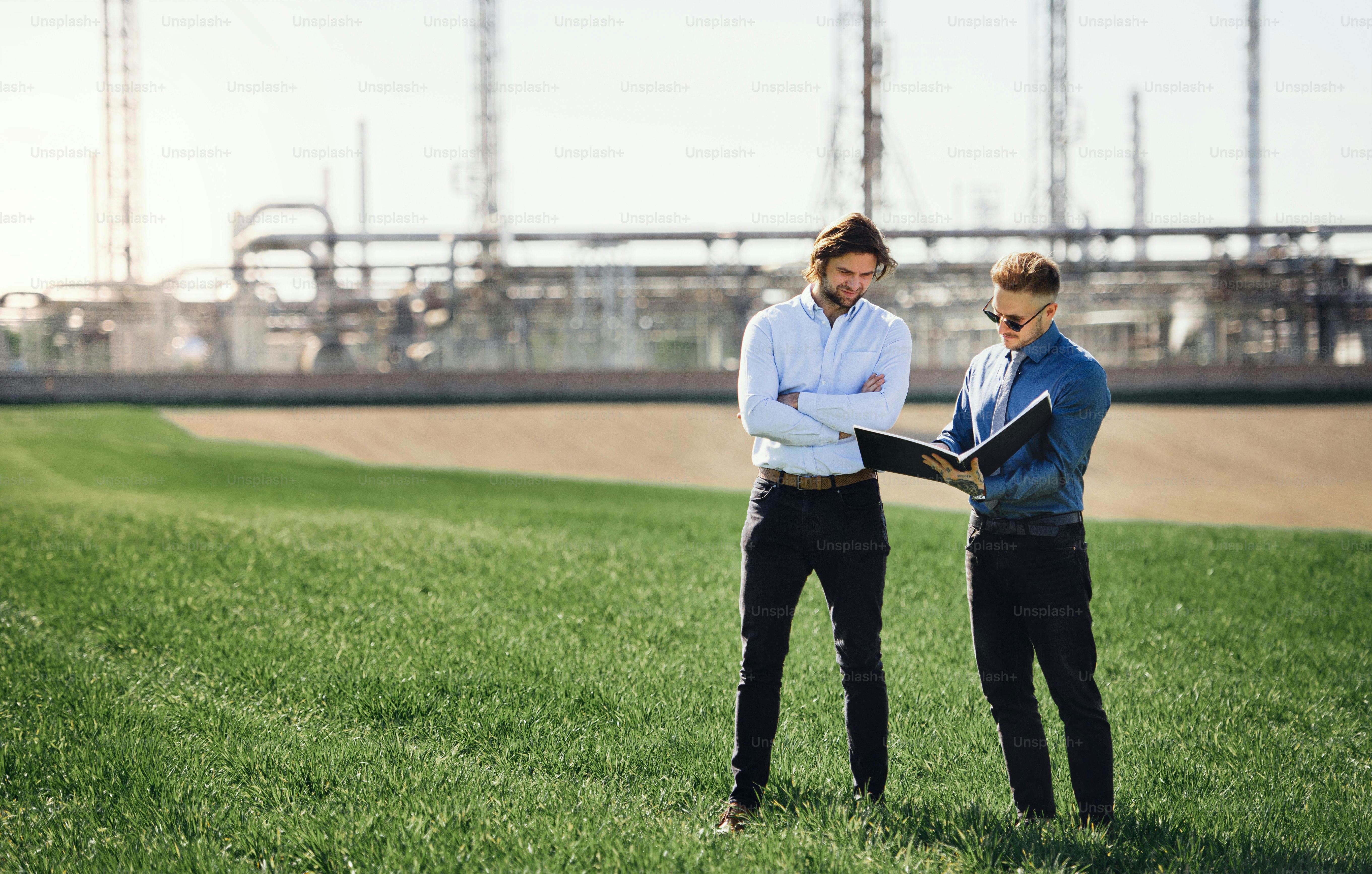 Two young engineers standing outdoors by oil refinery, discussing issues. Copy space.