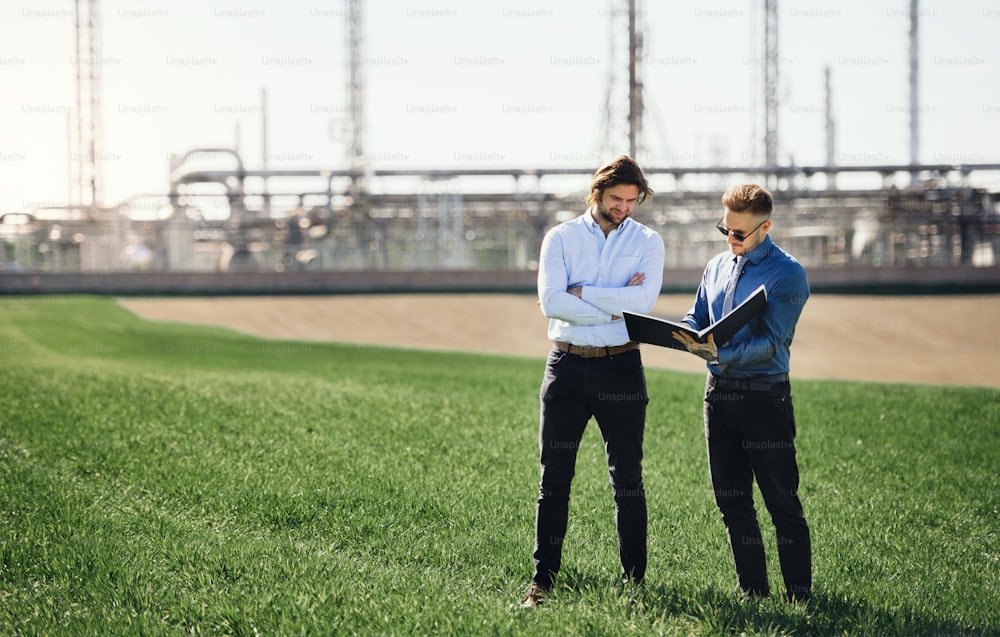 Two young engineers standing outdoors by oil refinery, discussing issues. Copy space.