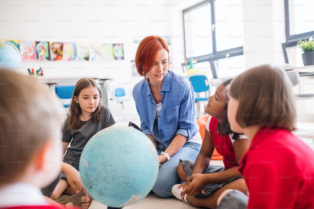 Un gruppo di piccoli scolari con un insegnante irriconoscibile seduto sul pavimento in classe, che impara la geografia.