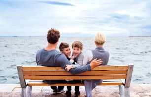 Young family with two small children sitting on bench outdoors on beach, rear view.