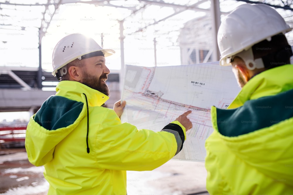 Rear view of men engineers standing outdoors on construction site, holding blueprints.