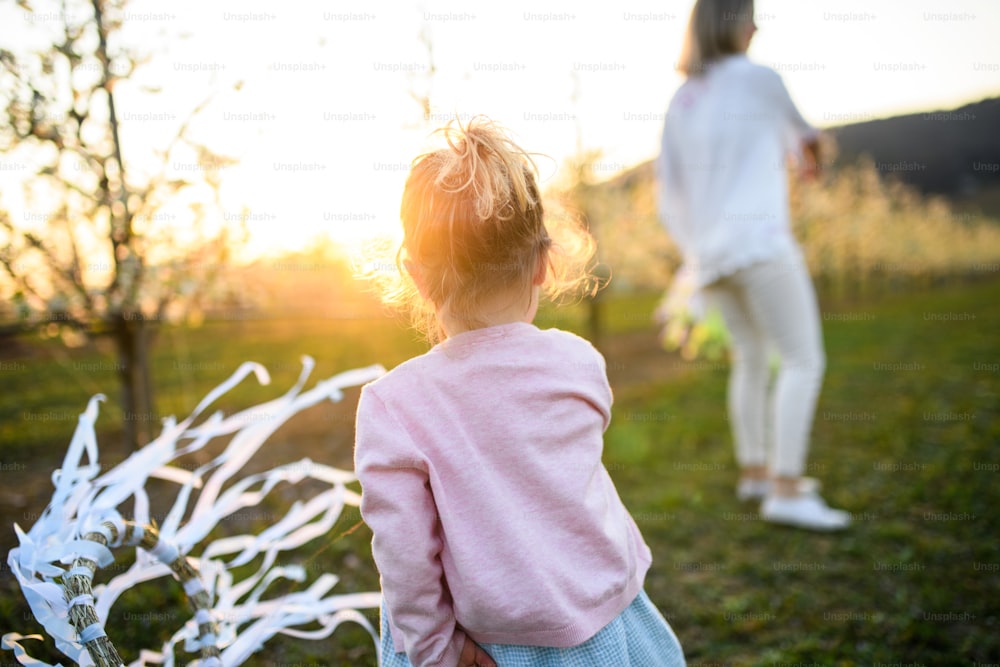 Rear view of small girl with unrecognizable mother playing outdoors in spring nature at sunset.