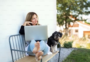Happy mature woman with dog working in home office outdoors on bench, using laptop.