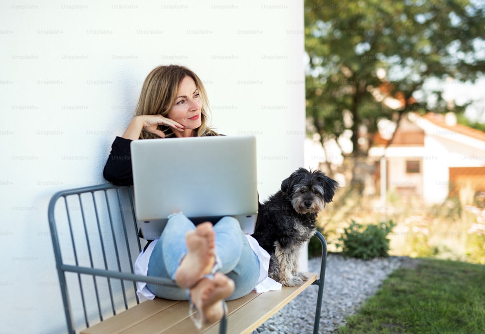 Happy mature woman with dog working in home office outdoors on bench, using laptop.