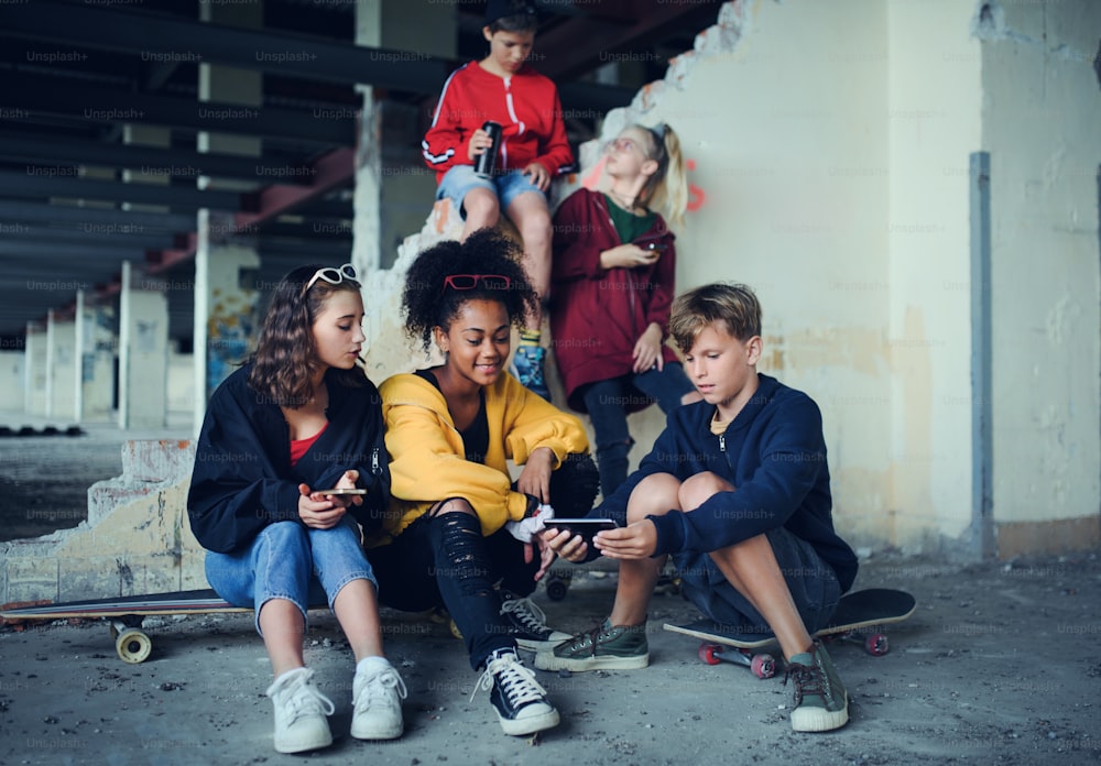 Front view of group of teenagers gang sitting indoors in abandoned building, using smartphones.
