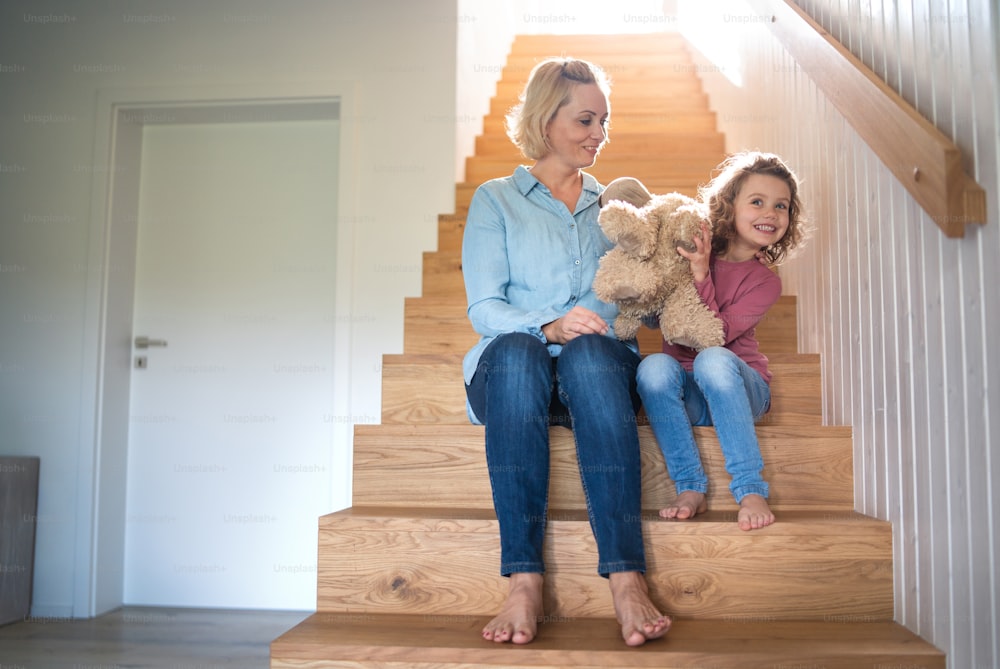 Eine Vorderansicht eines süßen kleinen Mädchens mit Mutter drinnen zu Hause, auf der Treppe sitzend.