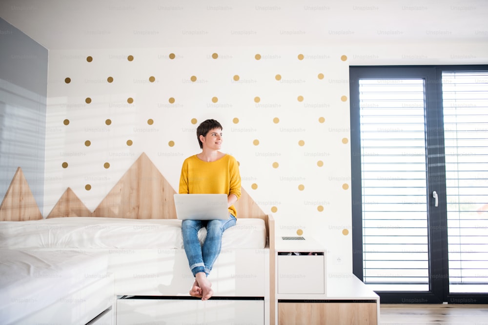 Front view of young woman with laptop sitting on bed in bedroom indoors at home.