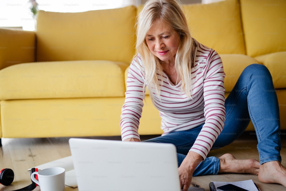 Senior woman with laptop indoors at home, relaxing on the floor.