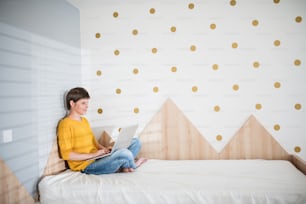 Side view of young woman with laptop sitting on bed in bedroom indoors at home.