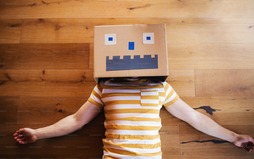 A top view of man with cardboard monster on head lying down on the floor indoors at home.