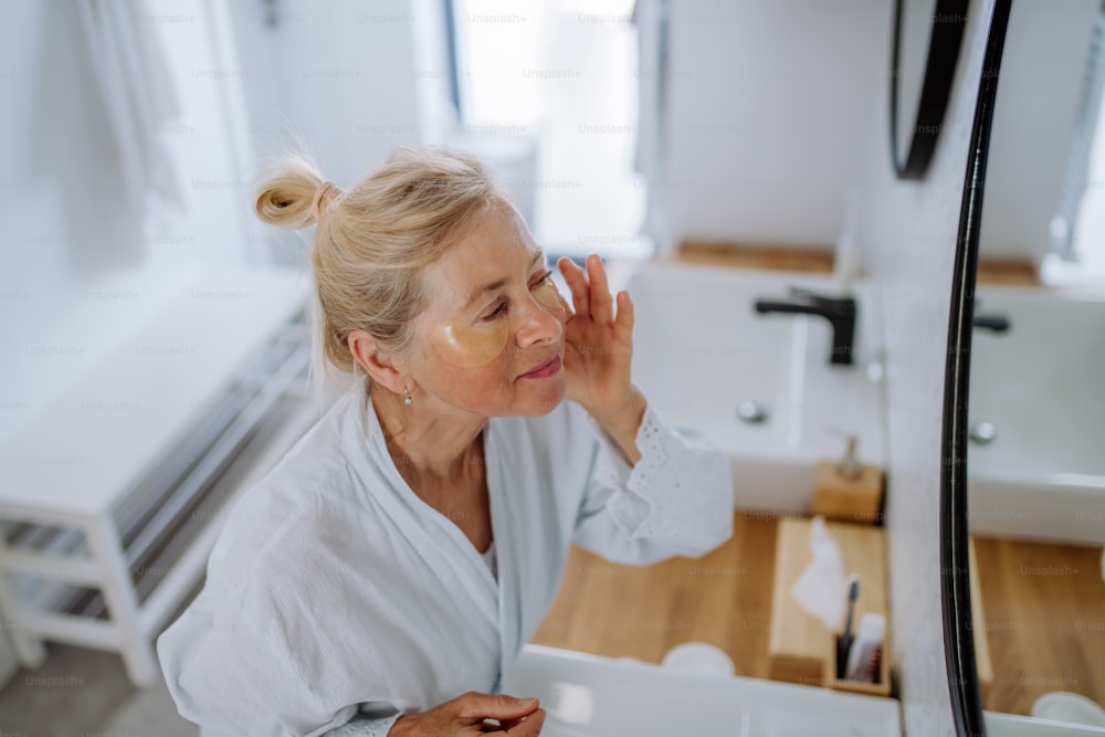 Eine schöne ältere Frau im Bademantel, die Augenklappen für Schwellungen aufträgt, während sie in den Spiegel schaut