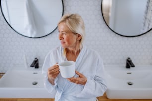 A beautiful senior woman in bathrobe drinking tea in bathroom, relax and wellness concept.