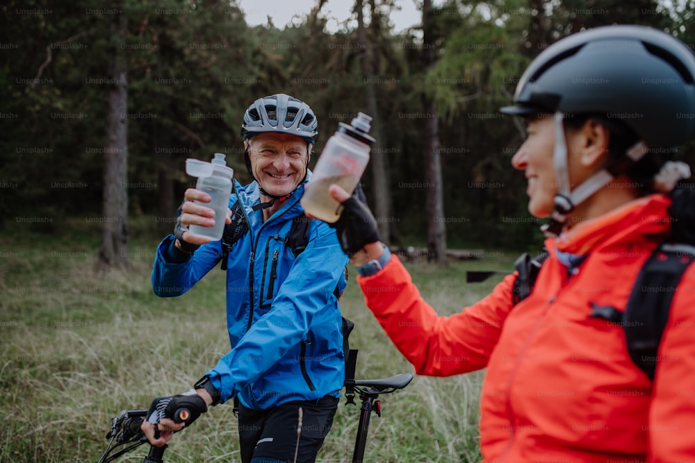 Una feliz pareja de ciclistas mayores con botellas de wtaer al aire libre en el bosque en un día de otoño.