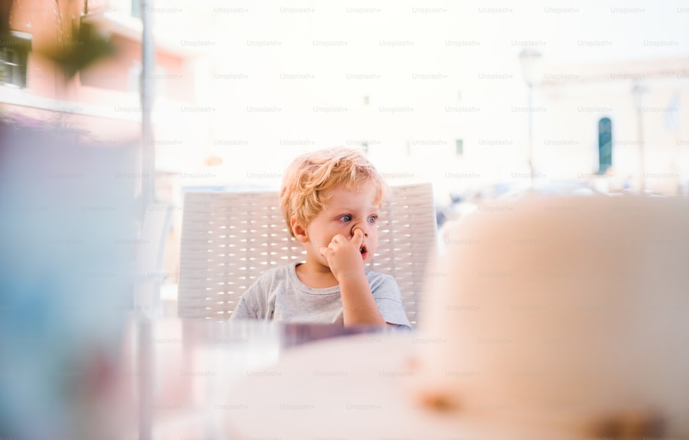 A small boy sitting in outdoor restaurant on summer holiday, picking nose.