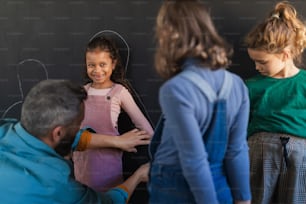 A teacher drawing little girl student with chalks on blackboard wall indoors in playroom.