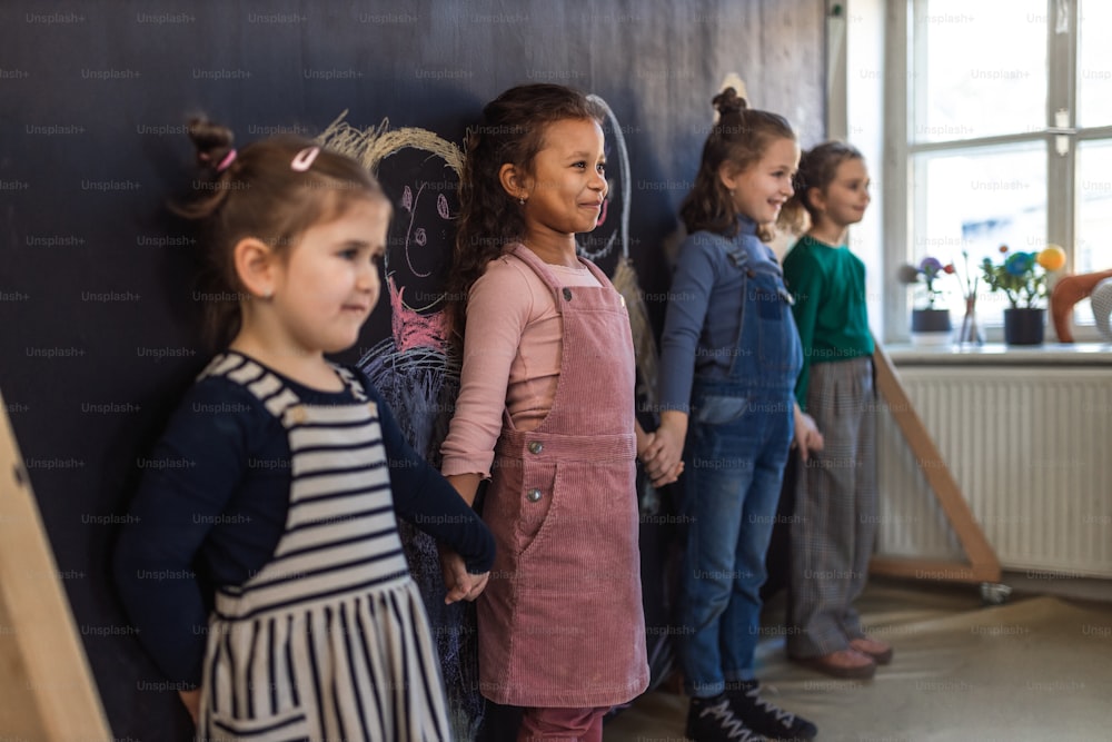 A group of little girls posing in front of blackboard wall paintings indoors in playroom.