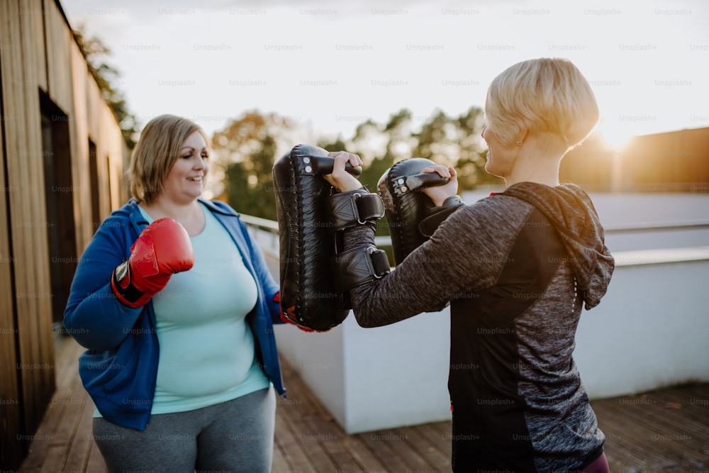 Una mujer con sobrepeso entrenando boxeo con entrenador personal al aire libre en terraza.