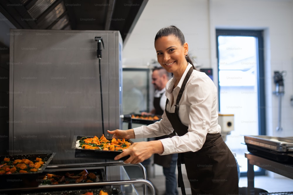A happy female cook looking at camera and holding tray with baked pumpkin pieces in commercial kitchen.