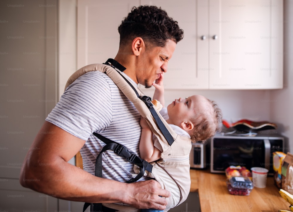 A father with small toddler son in carrier in kitchen indoors at home, having fun.