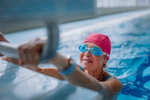 An active senior woman swimmer holding onto starting block preparing to swim in indoors swimming pool.