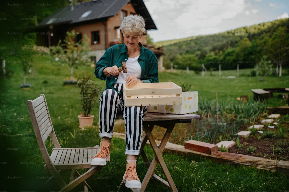A happy senior woman renovating wooden crate outdoors in garden.